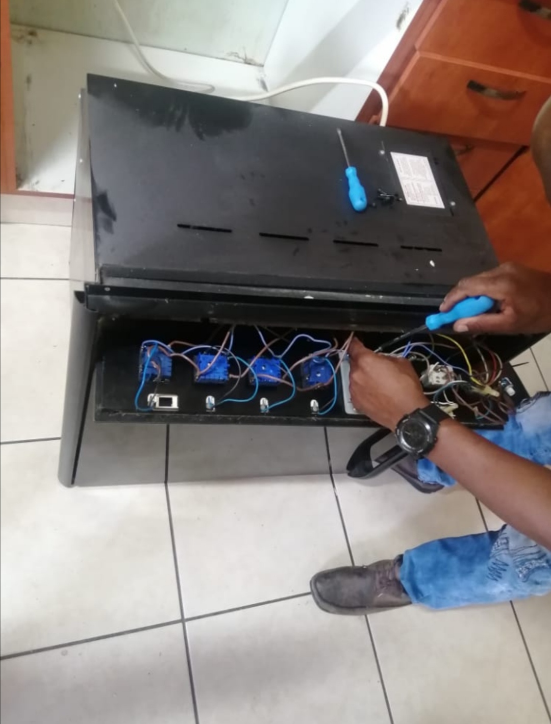 Underside of an oven with exposed wiring while a technician works on it with a screwdriver