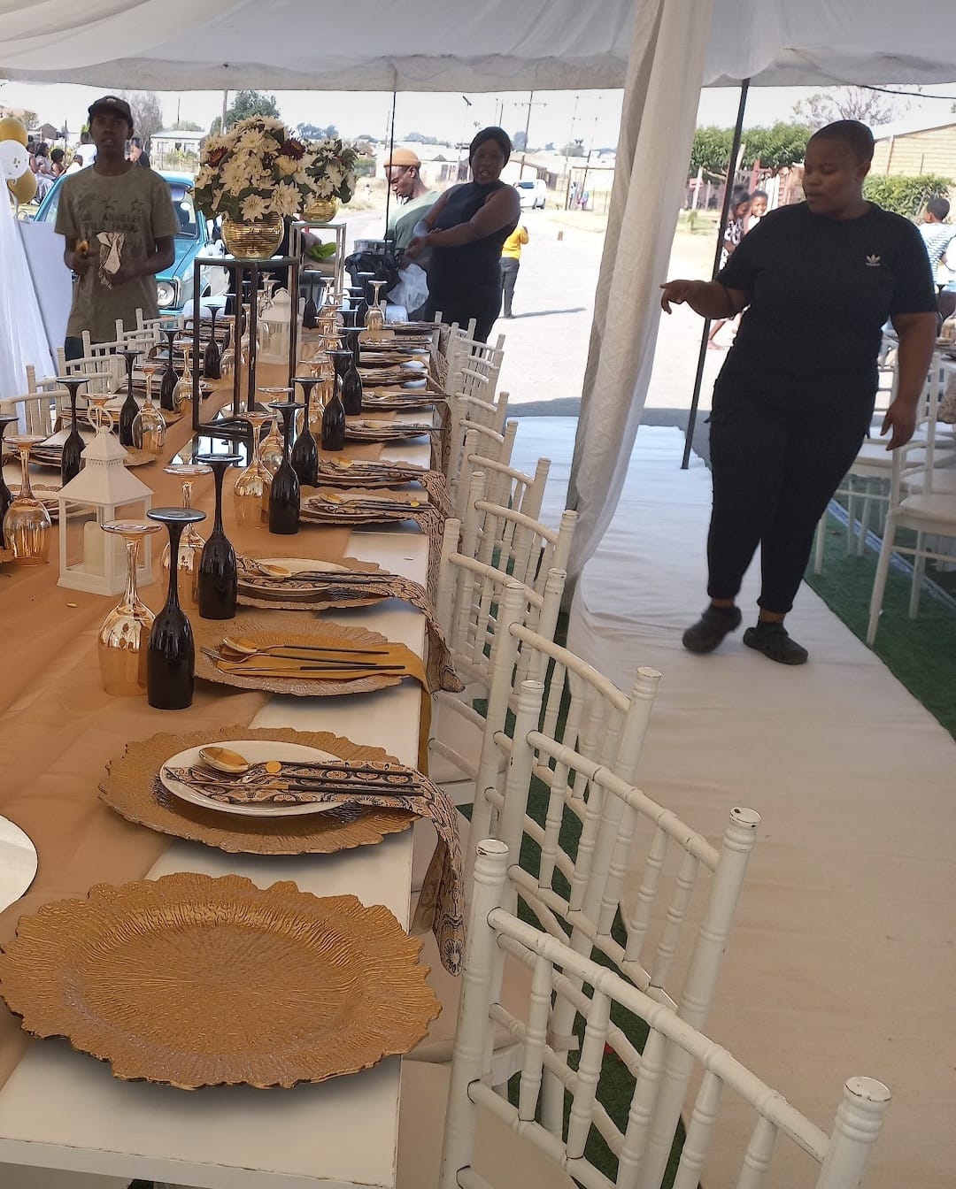 Long event table set with gold charger plates, black glassware and lanterns, with white chiavari chairs under a white marquee